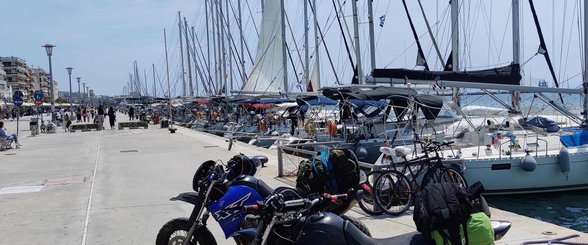 Auf einer Motorradtour durch Griechenland stehen ein schwarzes und ein blaues Motorrad mit Gepäck auf einem Kai mit einer Reihe von Segelbooten im Hintergrund an einem sonnigen Tag mit leicht bewölktem Himmel.