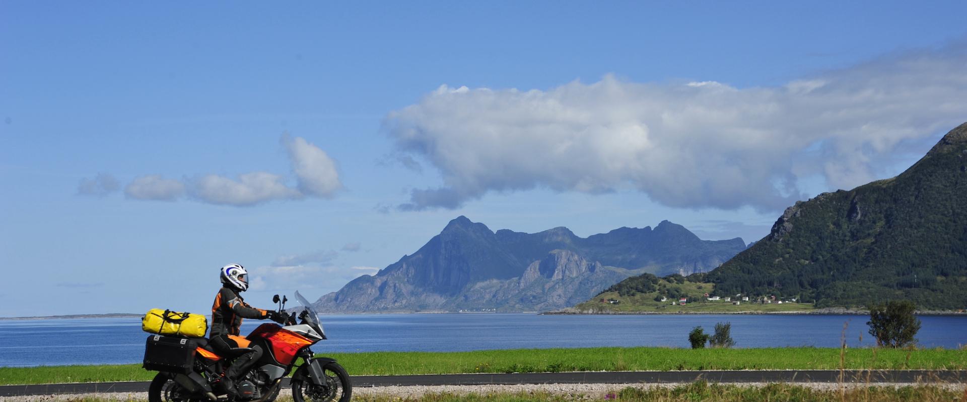 Motorrad mit gelben Koffern fährt auf einer Küstenstraße vor einer malerischen Bergkulisse und einem klaren blauen Himmel