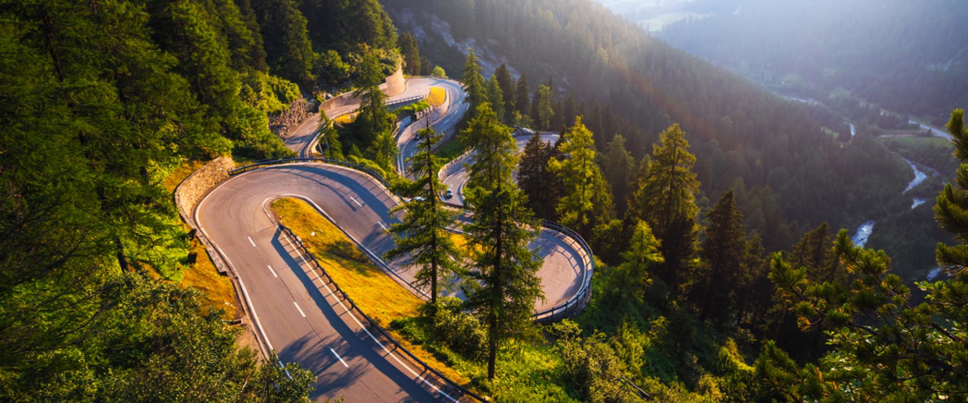 Der Maloja Pass, eine kurvige Bergstraße, windet sich berghoch durch einen dichten Wald, beleuchtet von warmem Sonnenlicht, mit einem Tal im Hintergrund.