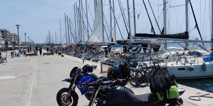 Auf einer Motorradtour durch Griechenland stehen ein schwarzes und ein blaues Motorrad mit Gepäck auf einem Kai mit einer Reihe von Segelbooten im Hintergrund an einem sonnigen Tag mit leicht bewölktem Himmel.