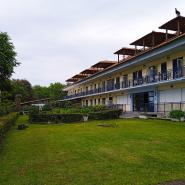Hotel Vlassis Stomio, gelb gestrichen mit Terrakotta-Dächern und blauen Geländern, vorne eine grüne Rasenfläche, eingebettet in eine grüne Landschaft unter einem bewölkten Himmel.
