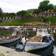 2 Fischerboote in einem kleinen Hafen, mit Netzen an Bord, im Hintergrund das Hotelgebäude Vlassis Stomio und eine bewaldete Hügellandschaft unter einem bedeckten Himmel.