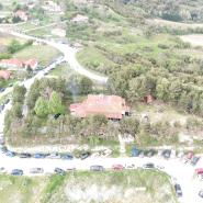 Luftaufnahme eines ländlichen Gebiets mit einem zentralen Gebäude, der Sikamines Olympus Lodge, umgeben von einem Parkplatz voller Autos, eingeschlossen von bewaldeten Gebieten und Feldern, mit einer Straße, die sich durch die Landschaft schlängelt.