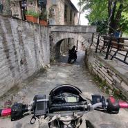 Blick aus der Fahrerperspektive eines Motorrades, das sich einem alten Steintunnel in Keramidi nähert, während ein Motorrad gerade durchfährt, rechts ist ein Zaun und links ein Haus zu sehen