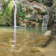 Ein kleiner, ruhiger Wasserfall fließt in einen klaren Naturteich, umgeben von felsigem Terrain und grüner Vegetation